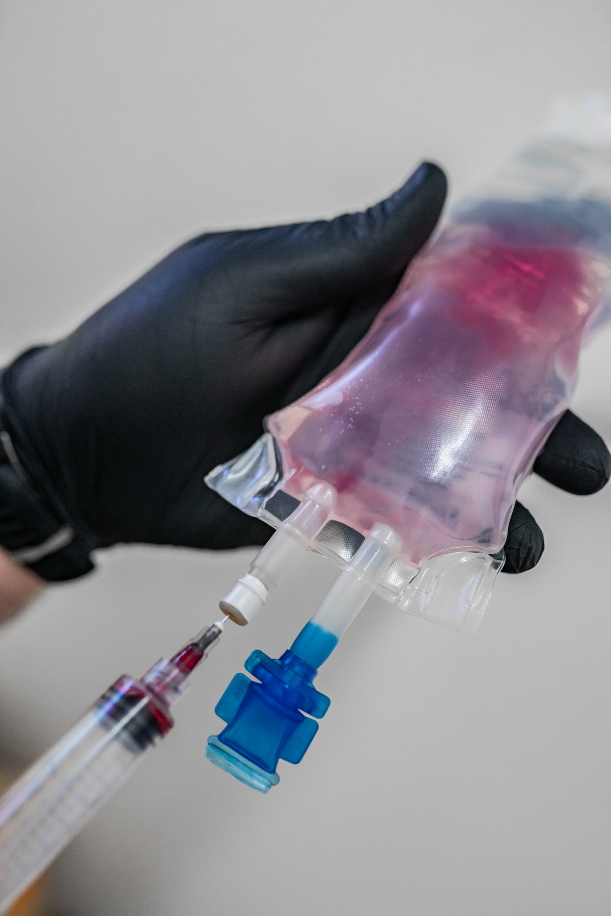 Hand with black glove holding a medical setup with a bag, needle, and syringe on a light background